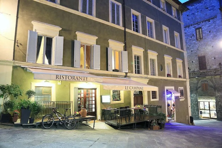 An evening exterior shot of a building housing "Ristorante Le Campane" with its illuminated awning and windows, featuring an outdoor seating area and a bicycle parked on the street in front.