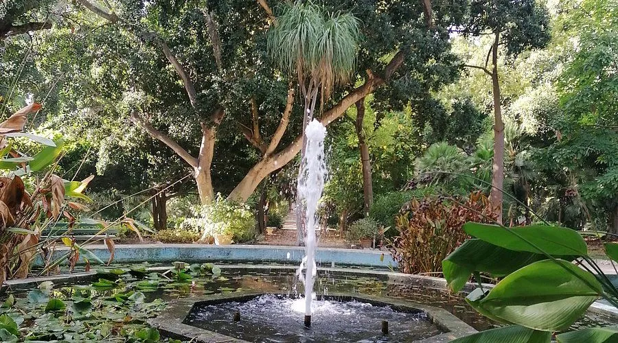 The image features a well-focused fountain as the central subject, set within a lush garden environment. The colors are natural and vibrant, and the lighting is generally even, contributing to a realistic depiction of the scene.