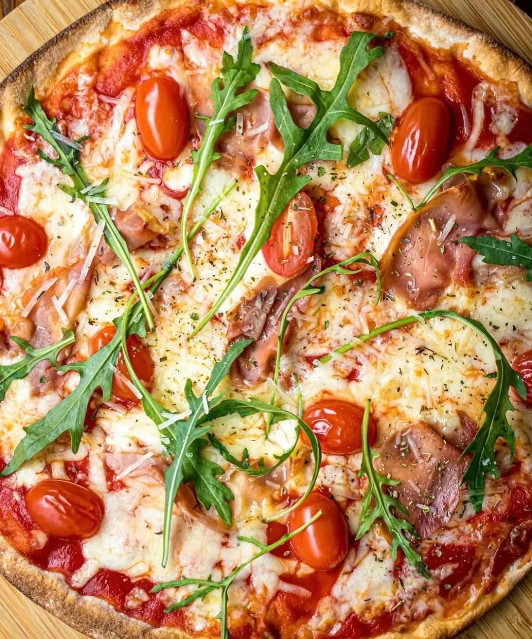 An overhead, close-up shot of a pizza topped with fresh arugula, cherry tomatoes, and thinly sliced ham. The cheese is melted and bubbly, and the crust shows a golden-brown edge. The ingredients are evenly distributed, creating an appetizing visual.
