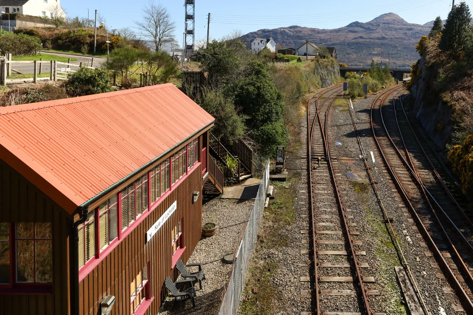 An outdoor photograph of a railway station building and multiple tracks, set against a backdrop of hills and a clear blue sky. The scene is well-lit by natural sunlight.