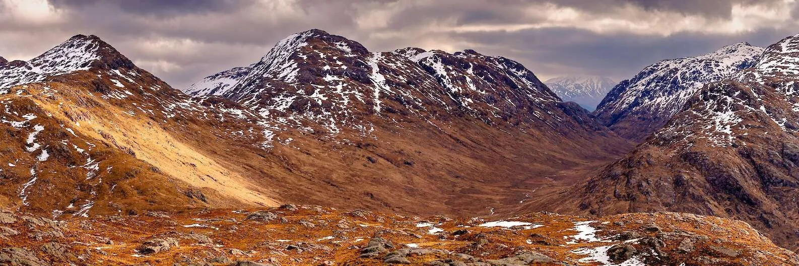 A rugged mountain landscape featuring a large, snow-dusted mountain under a cloudy sky, with a foreground of brown and reddish rocky terrain.