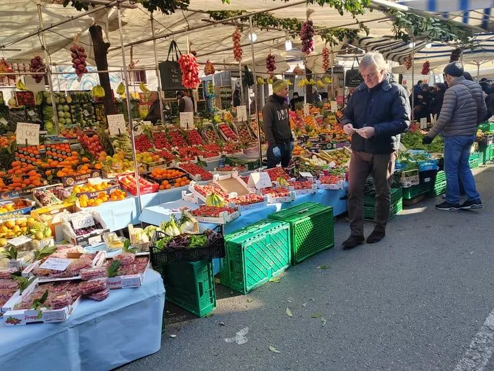 The image effectively captures a vibrant outdoor market scene with an abundance of fresh produce, showcasing good natural lighting and accurate, lively colors. The overall sharpness is commendable, with key elements like the fruits, vegetables, and individuals in the foreground and midground being in focus. The photo-realistic quality contributes significantly to its strength. Some minor weaknesses include a slightly less sharp background and a composition that, while capturing the essence of a market, could be considered busy.