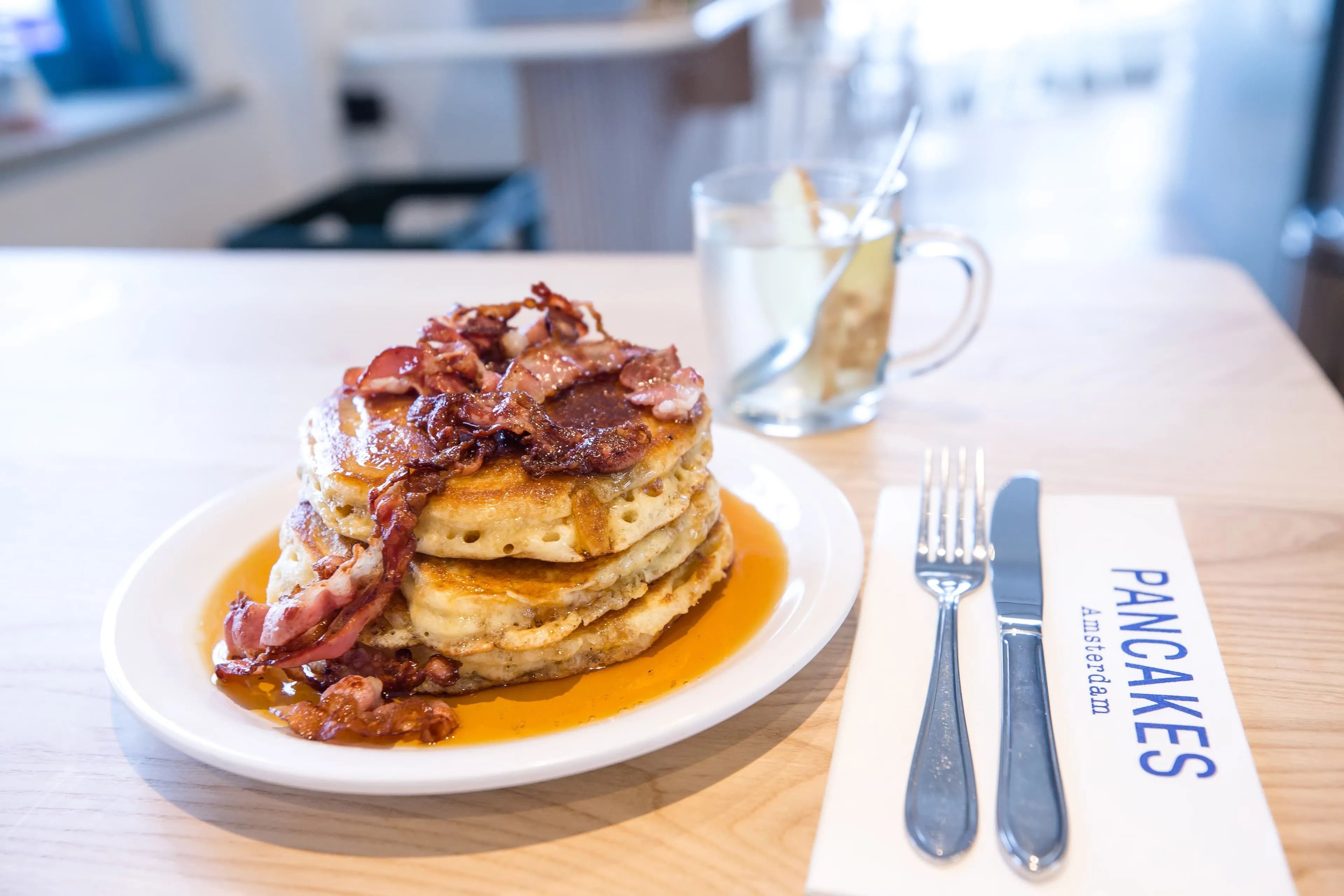 A well-composed and appetizing image featuring a stack of golden pancakes generously topped with crispy bacon and drizzled with maple syrup. The pancakes are the clear focus, with cutlery and a drink in the soft-focus background, suggesting a breakfast or brunch setting.