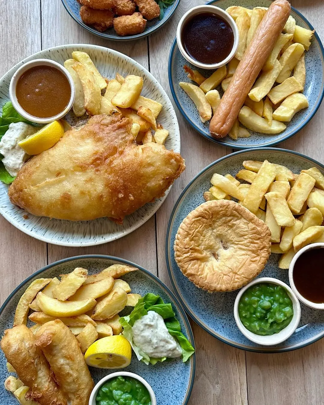 The image features multiple plates of classic British fare, including fish and chips, pie and chips with peas, and sausage and chips. The top-down composition effectively showcases all the dishes, which are well-lit with natural-looking light that highlights their textures. The image is sharp, demonstrating good focus on all key elements, and the colors are accurate and appealing, contributing to its high photo-realism.