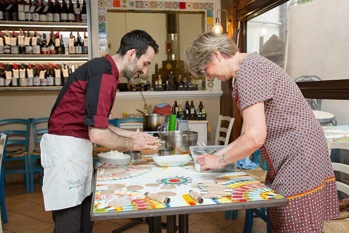 An image depicting two individuals, possibly a chef and a student, engaged in a cooking activity, likely making pasta or dough. They are focused on the task at a table with various cooking implements. The setting appears to be a professional kitchen or culinary workshop.