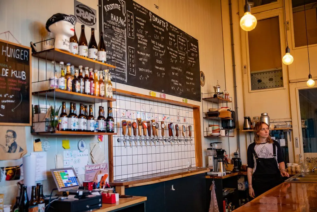 An inviting interior shot of a craft beer bar or brewery, featuring a prominent wall of tap handles, shelves stocked with bottled beverages, and a large chalkboard menu. The lighting is warm, contributing to a cozy ambiance.