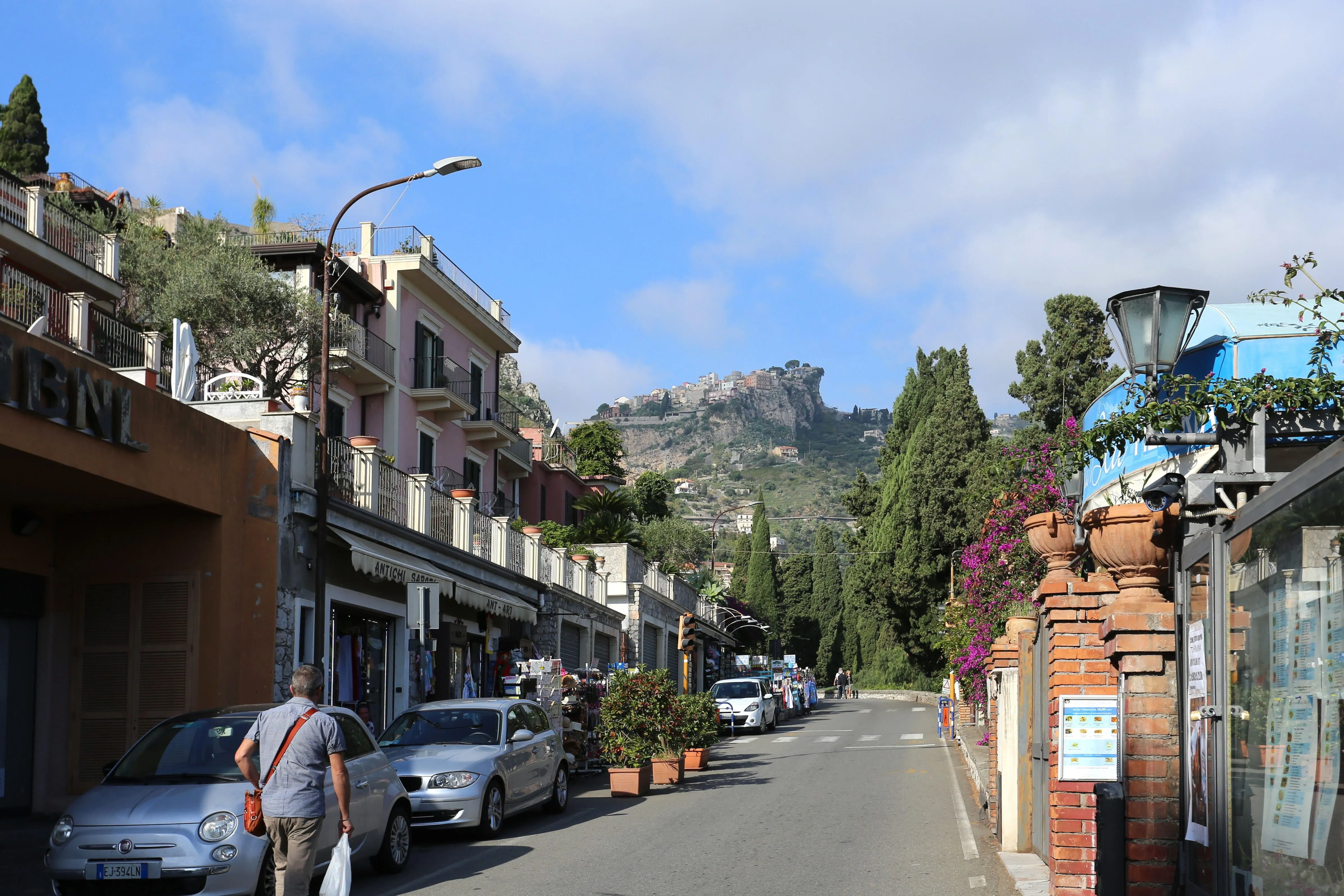 A street scene with buildings and parked cars lining the road, leading towards a distant town perched on a hill under a blue sky.