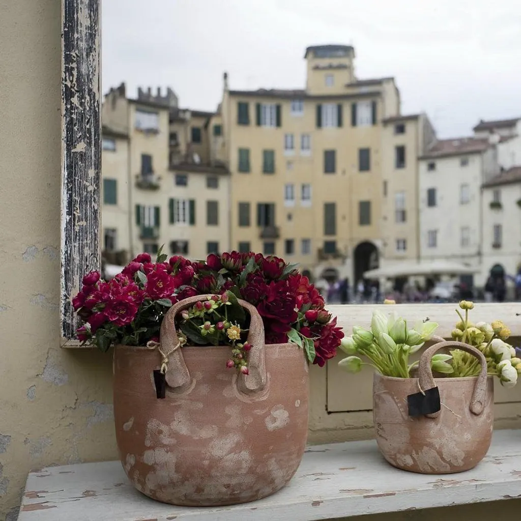 An editorial-style image featuring three terracotta pots filled with various flowers (red, green, and blue/purple) placed on a window ledge. The pots are in sharp focus, while the background depicts a blurred European city square with historical buildings and faint pedestrian activity, creating a sense of depth.