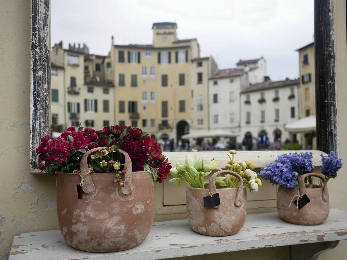 An editorial-style image featuring three terracotta pots filled with various flowers (red, green, and blue/purple) placed on a window ledge. The pots are in sharp focus, while the background depicts a blurred European city square with historical buildings and faint pedestrian activity, creating a sense of depth.