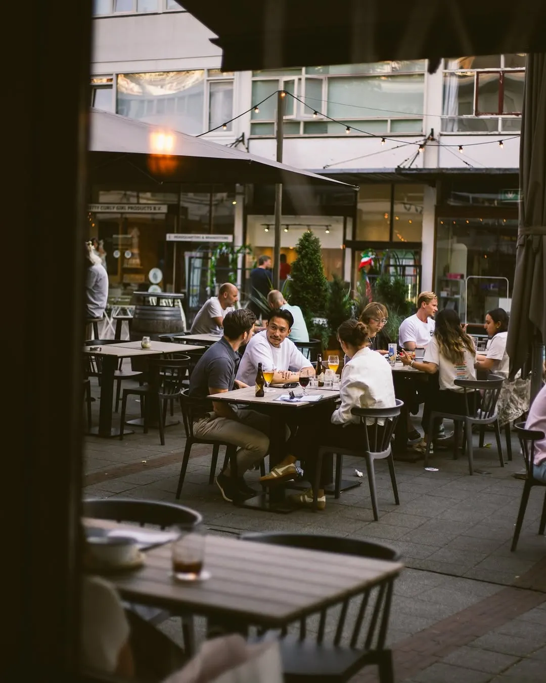 An inviting outdoor dining scene featuring several groups of people enjoying drinks and conversations at tables in what appears to be a city square or pedestrian area. The image has a warm, natural light and good depth of field.