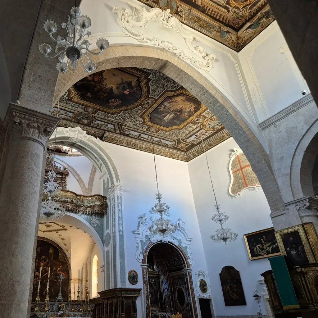 An interior view of a grand, ornate church or cathedral, featuring intricate ceiling paintings, decorative arches, large pillars, and elegant chandeliers. The architectural details are characteristic of a Baroque or similar historical style, showcasing rich artistry and craftsmanship.