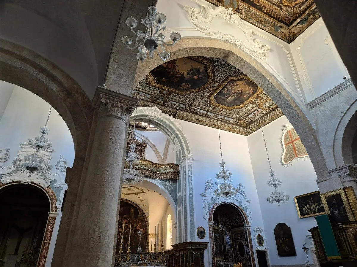 An interior view of a grand, ornate church or cathedral, featuring intricate ceiling paintings, decorative arches, large pillars, and elegant chandeliers. The architectural details are characteristic of a Baroque or similar historical style, showcasing rich artistry and craftsmanship.