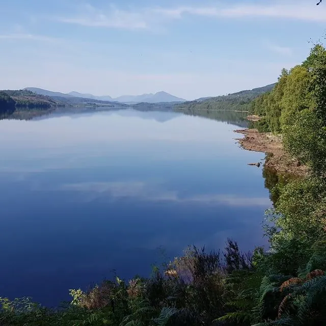 A serene landscape photograph featuring a calm body of water, likely a lake or reservoir, perfectly reflecting the blue sky and distant mountains. Lush green trees and foliage frame the right and foreground of the image.