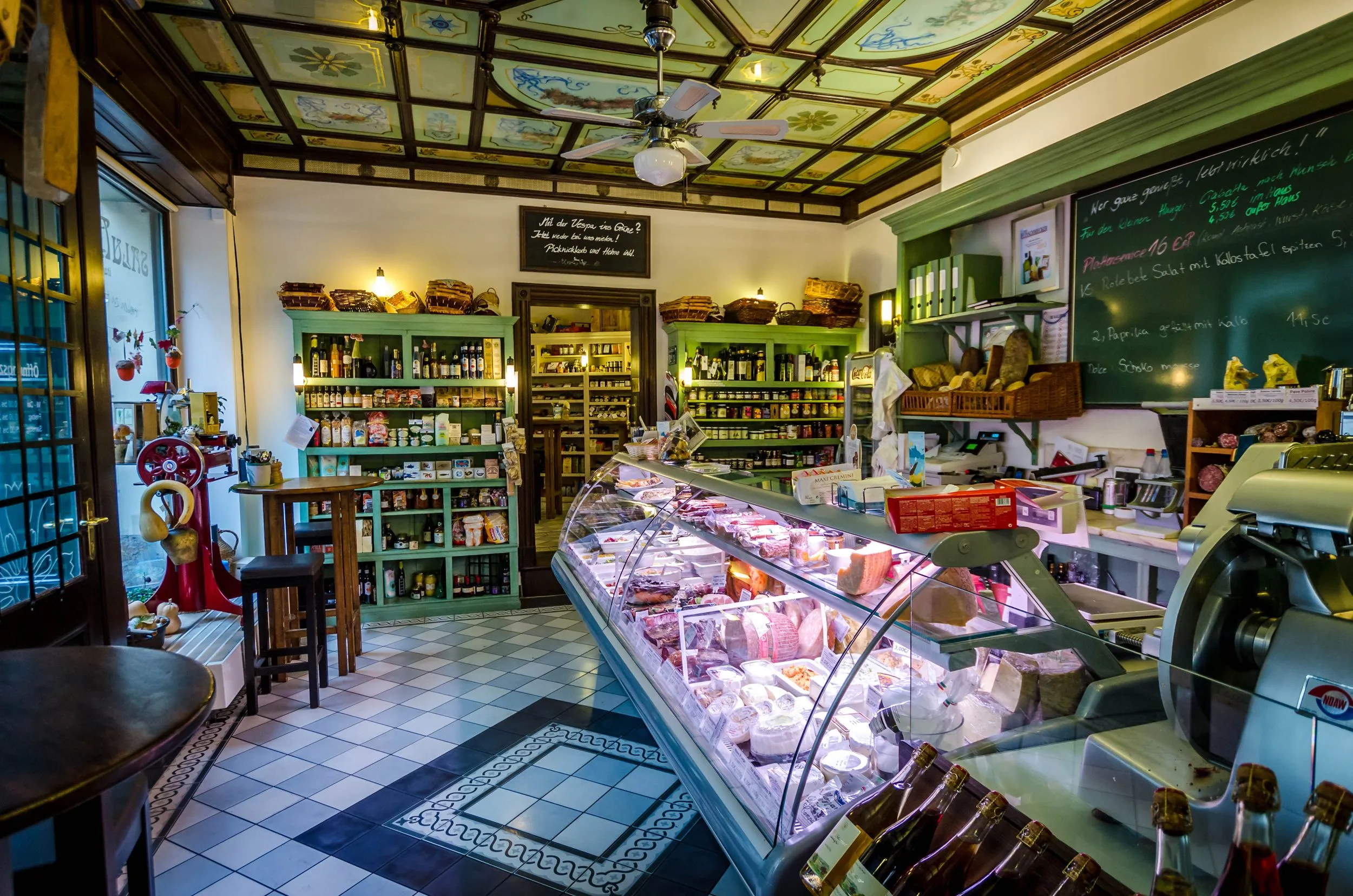 An interior shot of a traditional deli or food shop, featuring a large glass display counter filled with various food items like cheeses and cured meats. Shelves stocked with bottles and jars line the walls, and a decorative ceiling and tiled floor add character. The lighting is good, highlighting the products.