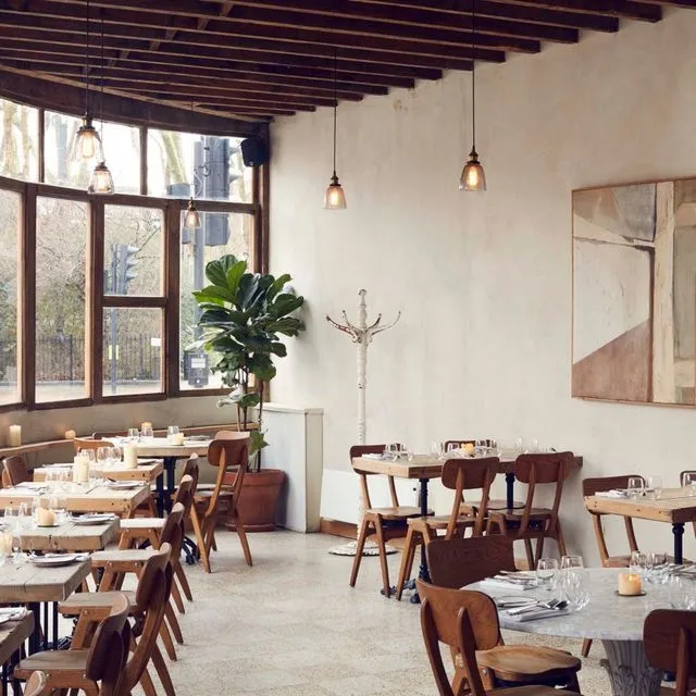 This image effectively showcases a modern restaurant interior with excellent natural lighting from large windows, creating a bright and inviting ambiance. The composition is well-balanced, highlighting the seating arrangements, wooden furniture, and decorative plant. The image is sharp and photo-realistic, with accurate colors. All key elements, such as the tables and chairs, are clearly in focus. There are no significant weaknesses.