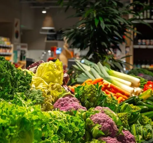 An editorial style image showcasing a vibrant and diverse selection of fresh vegetables, including various types of lettuce, purple and green cauliflower, carrots, and leeks, arranged appealingly on a display. The foreground is in sharp focus, while the background, depicting a blurred retail environment with ambient lighting, provides context.