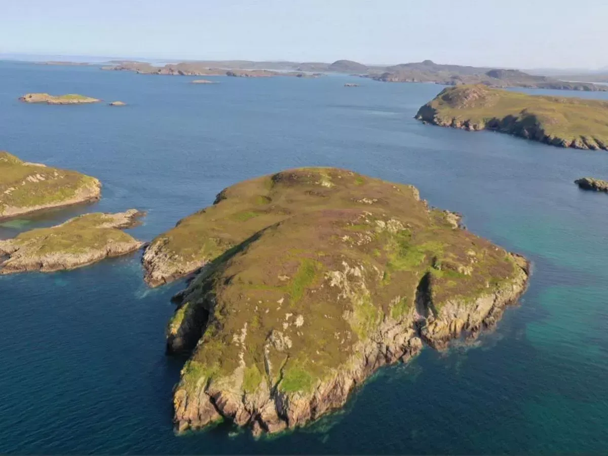 An aerial view of a small, green and brown island surrounded by clear blue water, with other landmasses visible in the distance under a bright sky.
