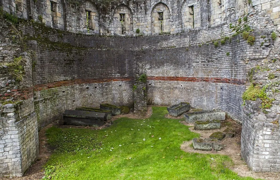 The image captures a historical stone structure, possibly a Roman amphitheater or bathhouse ruin, with grass covering the ground and ancient stone objects, resembling sarcophagi or troughs, scattered on the turf. The circular wall features several arched openings at a higher level, suggesting multiple stories or decorative elements. The overall scene conveys a sense of age and historical significance, with natural light illuminating the weathered stone and green vegetation.