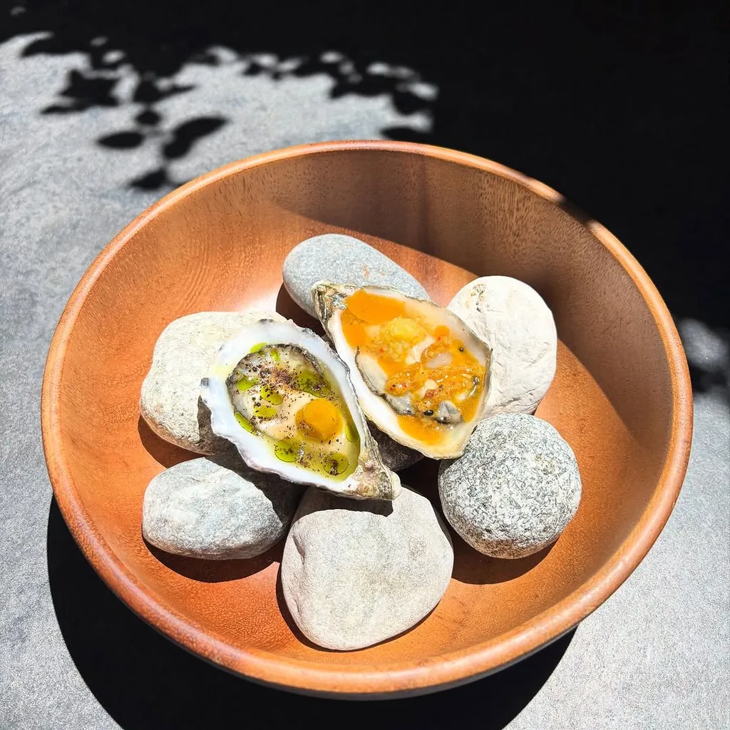 A professionally taken close-up image featuring two gourmet oysters, one adorned with green oil and the other with an orange sauce, presented artfully on natural stones inside a wooden bowl. The image captures the fresh and appetizing appearance of the dish.