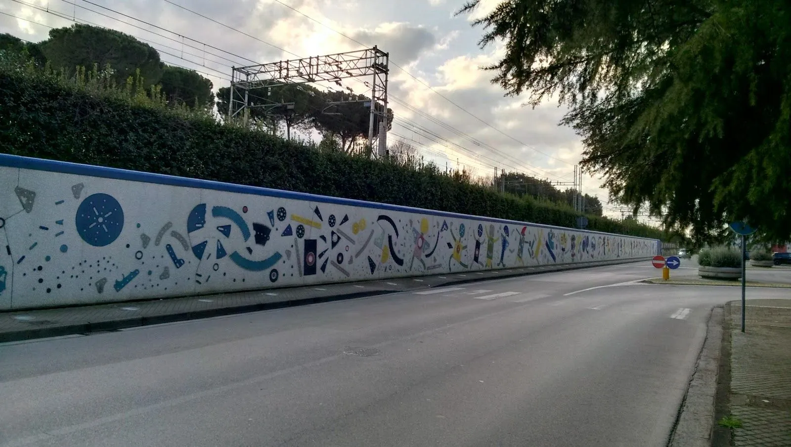 An outdoor shot of a long wall adorned with a colorful abstract mural, running alongside a road. In the background, there are trees and railway overhead structures beneath a cloudy sky.