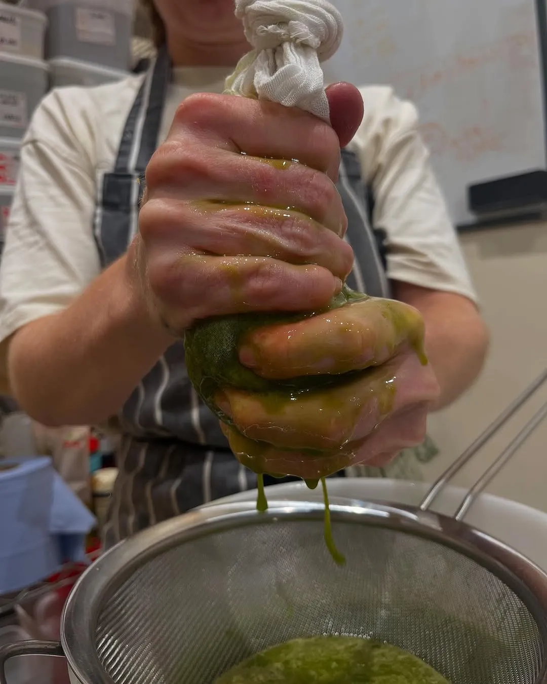 The image effectively captures the hands of a person squeezing a green substance into a strainer, depicting a food preparation process. The primary action and key elements, such as the hands and the dripping liquid, are sharp and well-focused. The lighting is sufficient, illuminating the scene clearly, and the colors appear natural and accurate. The image is highly photo-realistic, accurately representing the activity. However, the background is somewhat busy, slightly distracting from the main subject, and while well-lit, the lighting could be more dynamic for a higher score.
