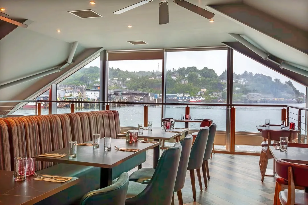 An image of a well-appointed restaurant interior featuring set tables and large windows offering a panoramic view of a waterfront town and hills. The dining area includes booth seating and individual chairs, providing a clear and inviting atmosphere.