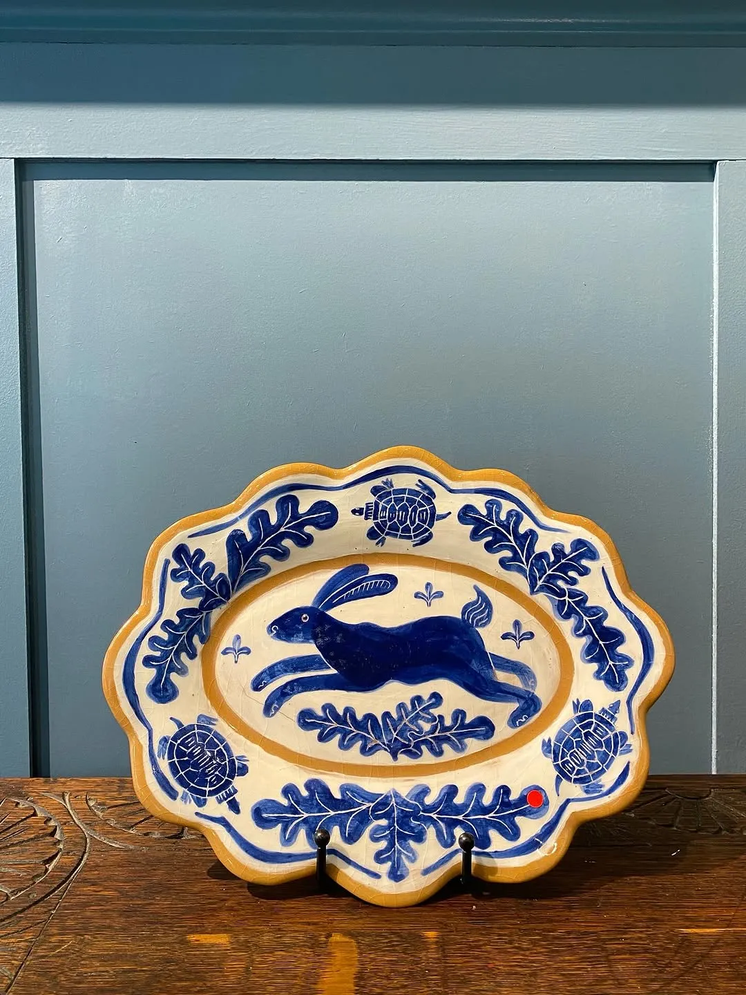 A well-composed photograph of a ceramic decorative plate featuring a blue rabbit and leaf pattern, presented on a wooden surface against a blue wall.