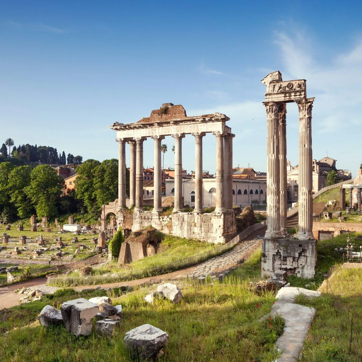 An outdoor daytime shot of the ancient ruins of the Roman Forum, featuring the Temple of Saturn and other remaining columns under a clear blue sky.