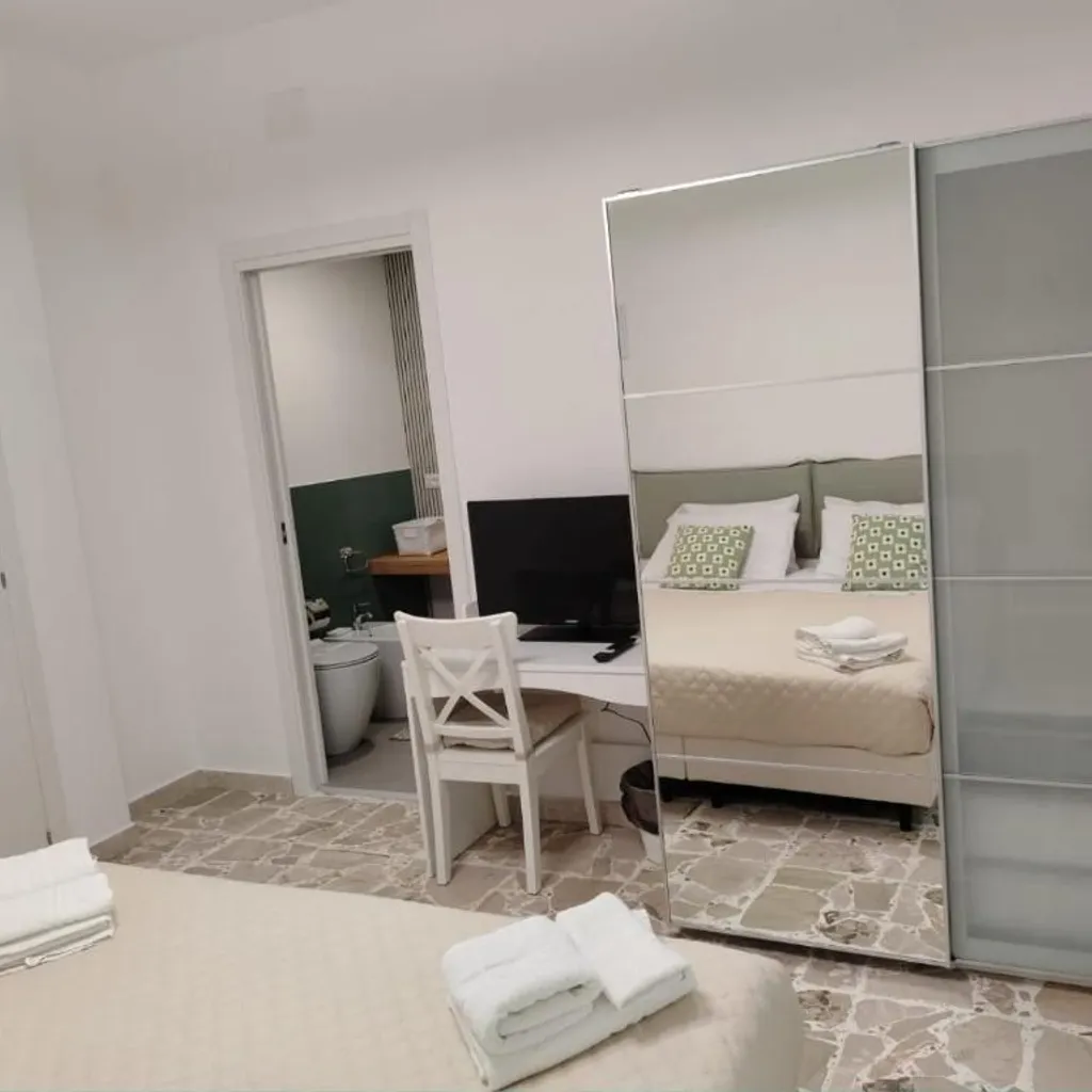 A view inside a clean and modern bedroom, likely in a hotel or guesthouse, featuring a bed with neatly folded white towels in the foreground. A desk with a television is visible on the left, next to a doorway leading to a bathroom. A large mirrored wardrobe reflects the bed and pillows, contributing to the sense of space. The room features light-colored walls and a patterned stone floor.