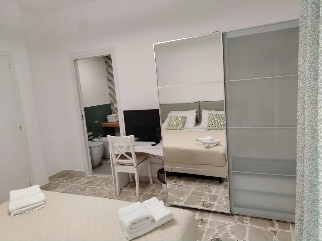 A view inside a clean and modern bedroom, likely in a hotel or guesthouse, featuring a bed with neatly folded white towels in the foreground. A desk with a television is visible on the left, next to a doorway leading to a bathroom. A large mirrored wardrobe reflects the bed and pillows, contributing to the sense of space. The room features light-colored walls and a patterned stone floor.