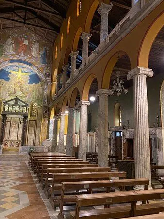 An editorial style image depicting the interior of a historic church or basilica, featuring rows of wooden pews, ornate marble columns, and richly decorated arches leading towards a detailed altar and mosaic-covered apse.