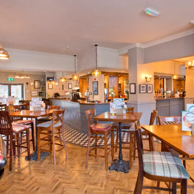 An inviting interior shot of a pub or restaurant, featuring wooden tables and chairs, a patterned tile floor near the bar, and warm lighting. The space appears clean and ready for patrons.