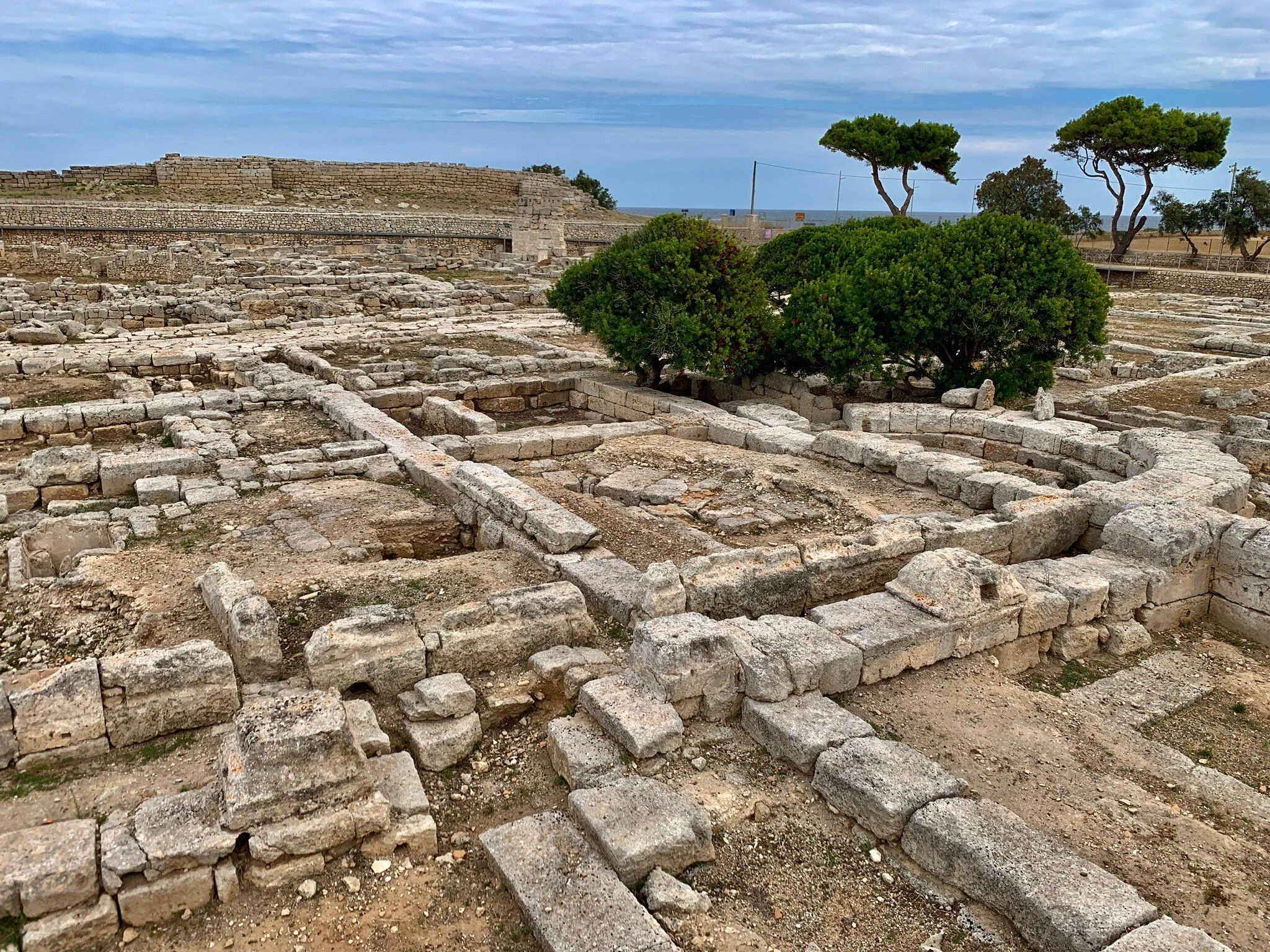 An archaeological site featuring ancient stone ruins and foundations, with green vegetation and the sea visible in the distance under a cloudy sky.