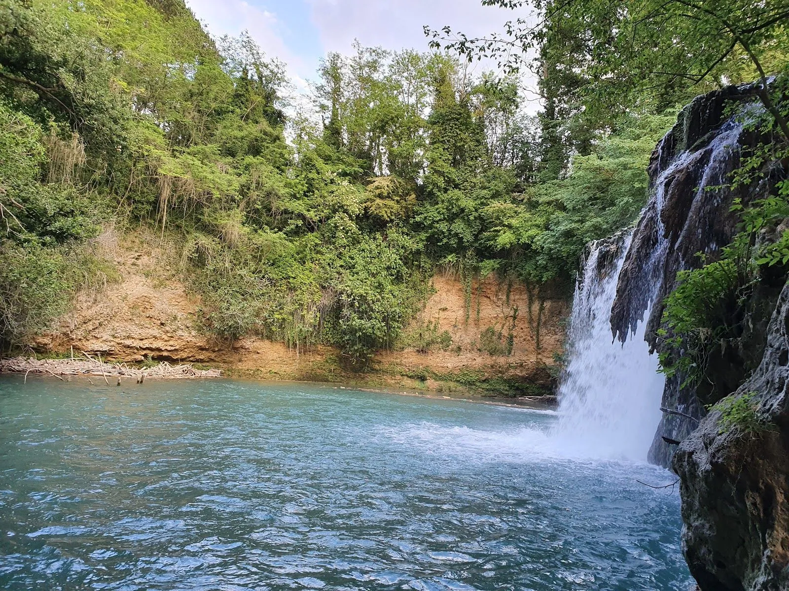 An editorial-style image capturing a picturesque waterfall cascading into a serene blue pool, surrounded by lush green foliage and a natural rock face.