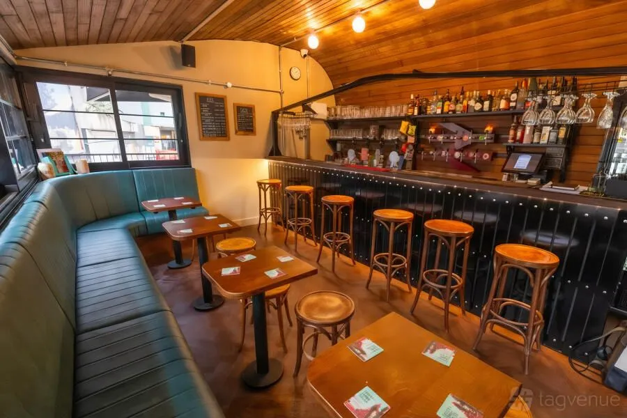 An image showcasing the interior of a bar or pub with a long bar counter, numerous bar stools, and booth seating with small tables. The space features warm wooden ceilings and flooring, complemented by ambient lighting and green upholstered seating.