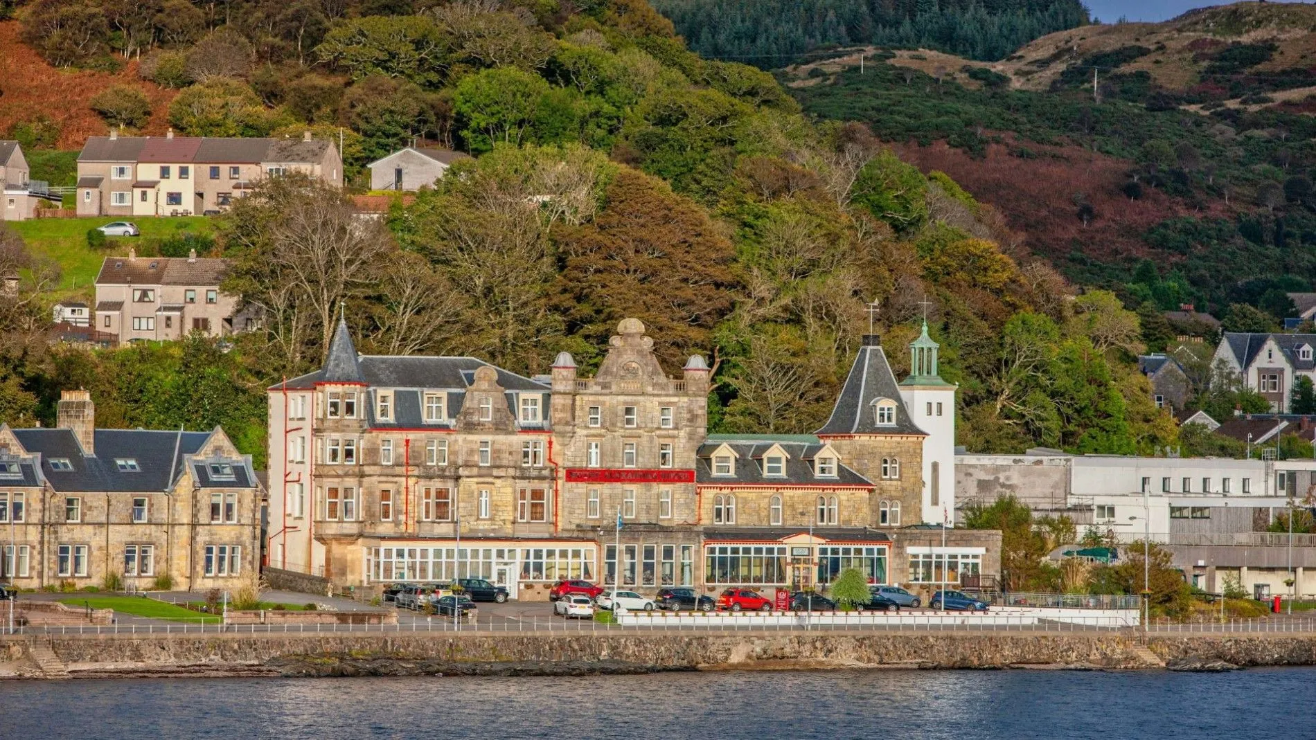 A large, classic stone building, likely a hotel, is situated along a waterfront with a tree-covered hillside rising steeply behind it. Several cars are parked in front of the building, which features distinct architectural styles including turrets and gabled roofs.
