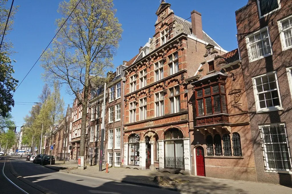 An outdoor shot showcasing a row of traditional European brick buildings lining a street on a clear, sunny day. A large tree with nascent leaves stands to the left, partially obscuring some of the structures. The architecture features ornate details, multiple windows, and gabled roofs, with vibrant red brickwork under a bright blue sky.