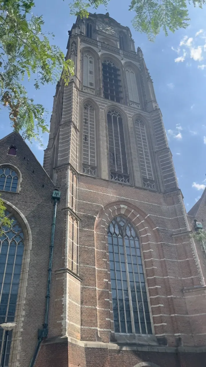 An outdoor photograph of a tall, historic church or cathedral tower constructed from brick and stone, featuring numerous large arched windows. The structure is set against a bright blue sky with scattered white clouds, with tree branches partially visible in the upper left of the frame.