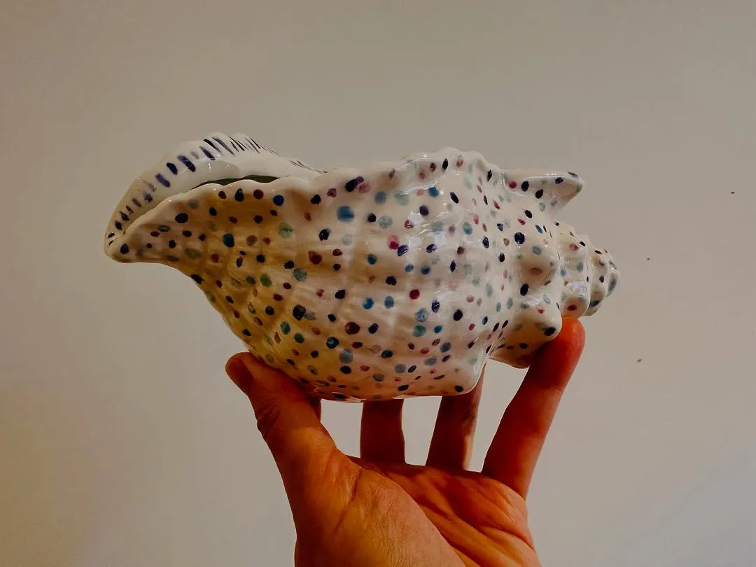 The image features a ceramic shell-shaped object with a multicolored polka-dot pattern, held in a person's hand. The main subject, the shell, is sharp and in clear focus, making the detailed pattern easily discernible. The composition effectively centers the object. However, the lighting is somewhat uneven, leading to some shadows on the hand and a slightly warm cast to the overall image, which impacts perfect color accuracy.