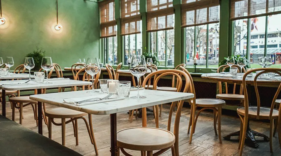 The image showcases a warm and inviting restaurant interior, well-lit by natural light from large windows. The foreground features a neatly set marble table with glasses and cutlery in sharp focus, while the background gracefully blurs. The green walls and wooden furniture contribute to a pleasant ambiance. The image is photo-realistic and colors appear accurate.