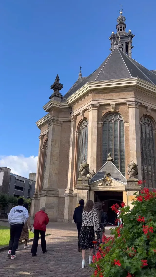 An outdoor shot of a historical building, likely a church or cathedral, featuring intricate architectural details under a clear blue sky. Several people are visible from behind, seemingly entering the building. Red flowers are present in the lower right corner, adding a touch of color.
