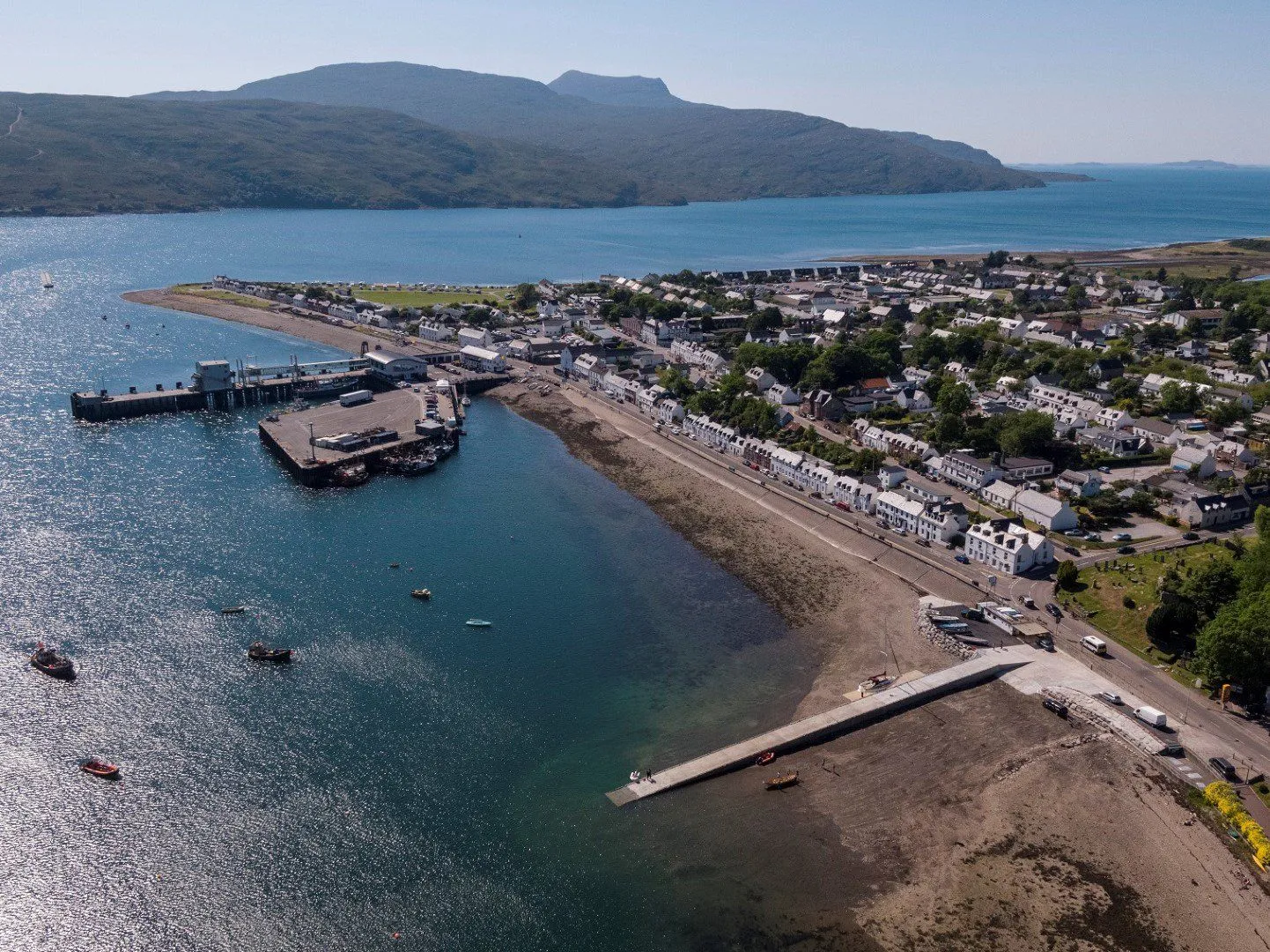 The image is an excellent aerial view of a coastal town, displaying strong composition and vibrant, accurate colors under good lighting. The entire scene is sharp and realistic, with all key elements, including the town, harbor, water, and distant mountains, in clear focus.