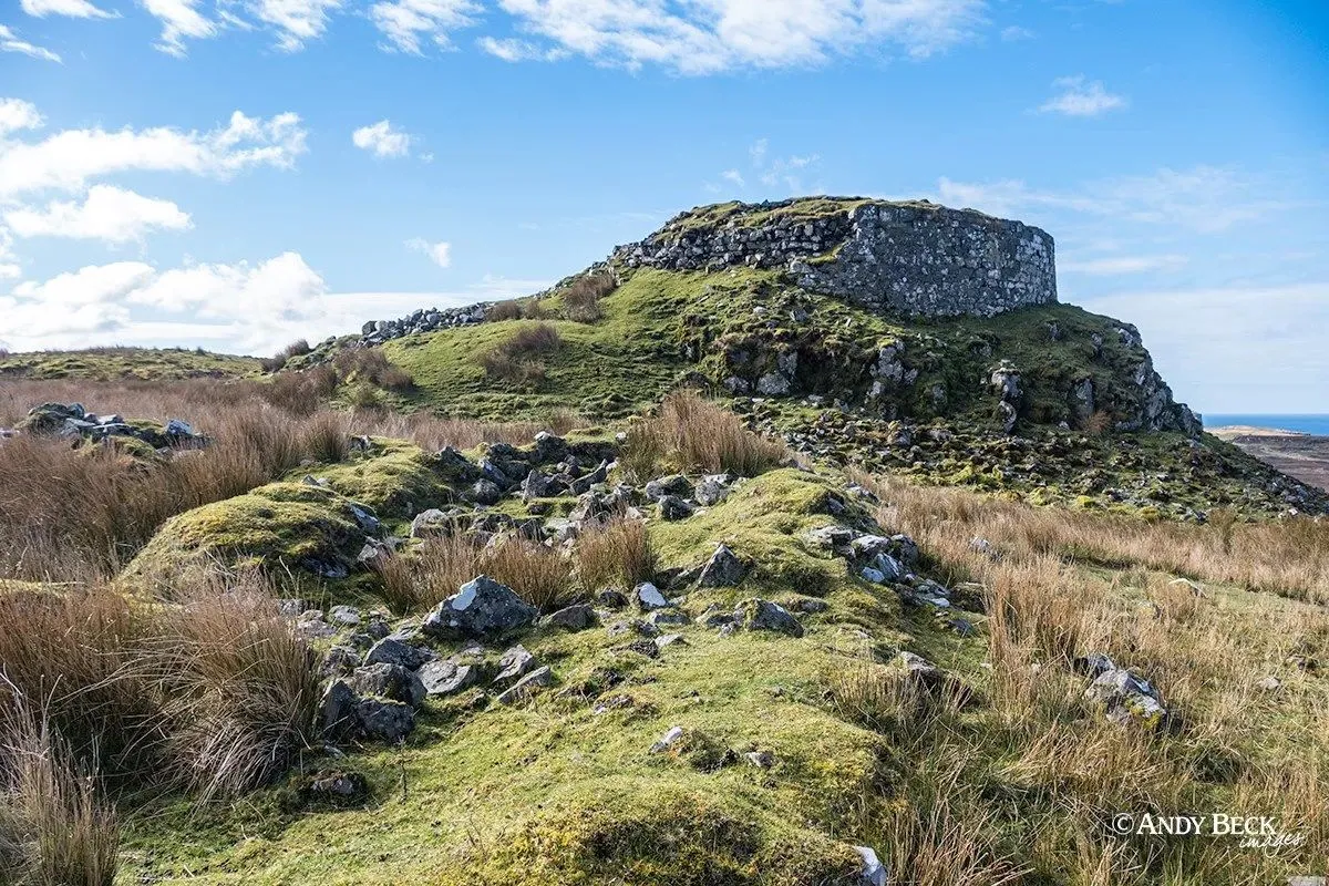 A well-composed and sharp photograph of an ancient stone structure on a green, rocky hill under a bright blue sky. The scene is well-lit with natural light, showing vibrant and accurate colors.