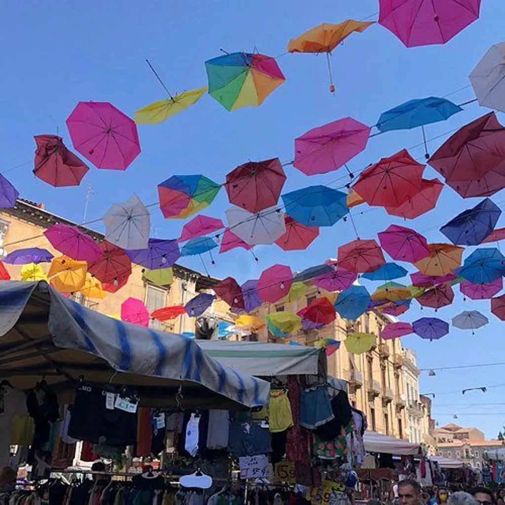 An outdoor market scene where numerous colorful umbrellas are suspended overhead, forming a vibrant canopy against a clear blue sky. Below, market stalls with various goods are visible, giving a sense of a lively public space.