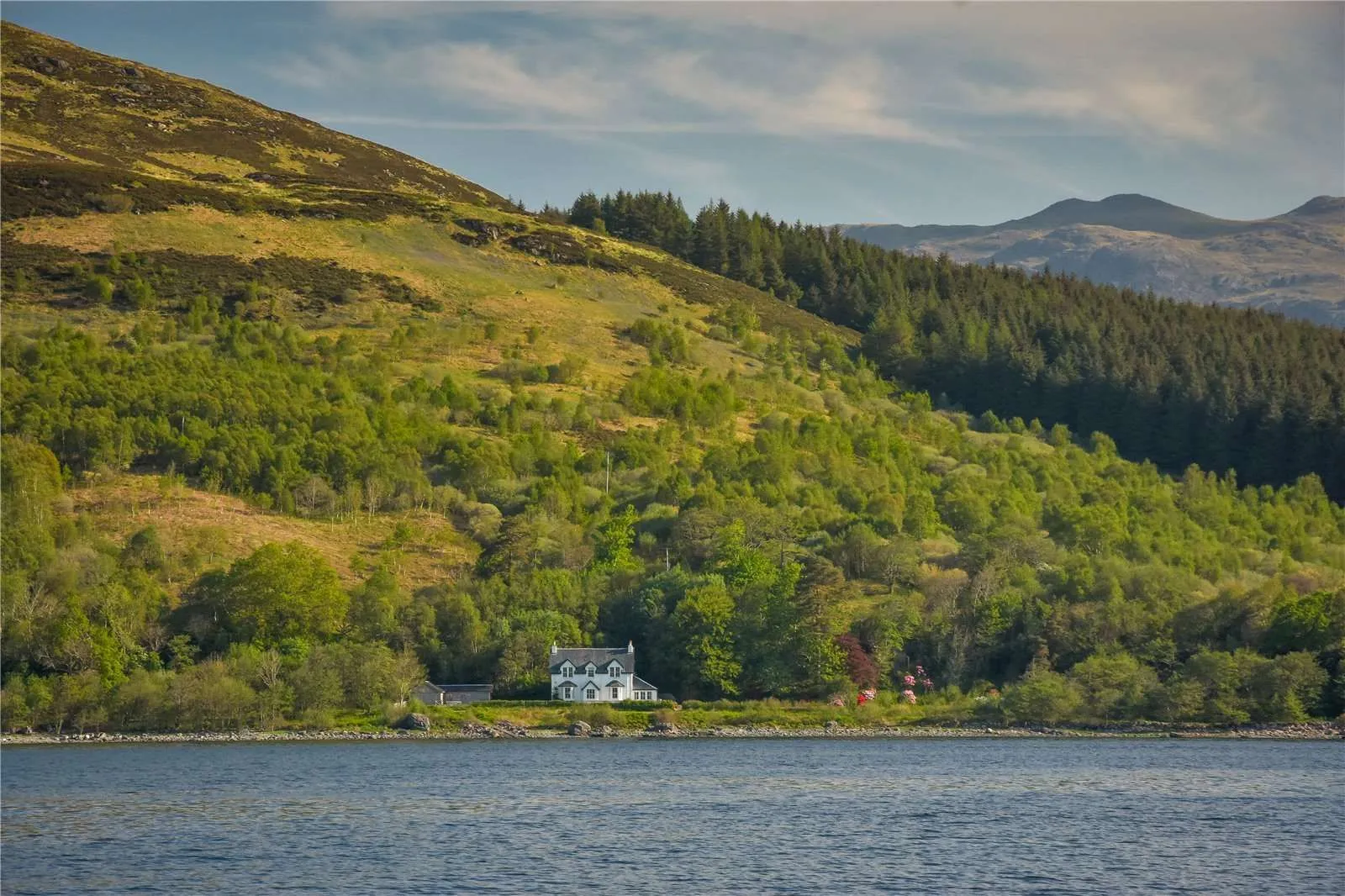 A picturesque white house stands by the tranquil water's edge, nestled at the base of a verdant, tree-covered hillside under a partly cloudy sky.