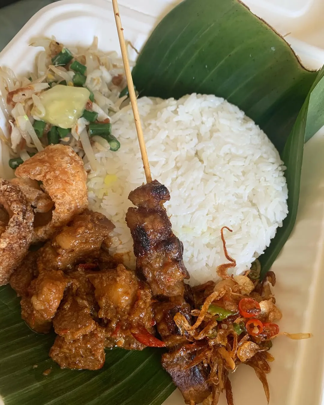The image features a well-composed and brightly lit close-up of a food platter, showcasing various elements including rice, a meat skewer, a stew-like dish, and vegetables, all presented on a banana leaf. The colors are natural and accurate, and the key food items are sharp and in focus, contributing to a highly photo-realistic appearance.