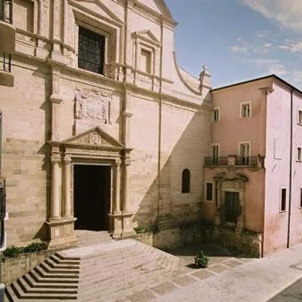 An outdoor architectural shot featuring a light-colored, ornate church facade on the left and a multi-story pink building on the right, with a street receding into the distance under a partly cloudy blue sky.