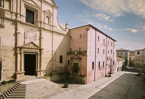 An outdoor architectural shot featuring a light-colored, ornate church facade on the left and a multi-story pink building on the right, with a street receding into the distance under a partly cloudy blue sky.