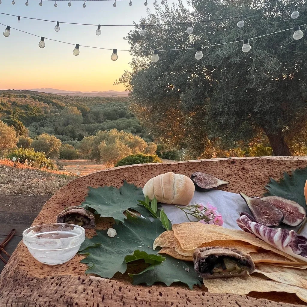 An inviting outdoor dining setup featuring a rustic table with food and a drink bowl, set against a stunning sunset landscape with rolling hills, olive trees, and string lights overhead.