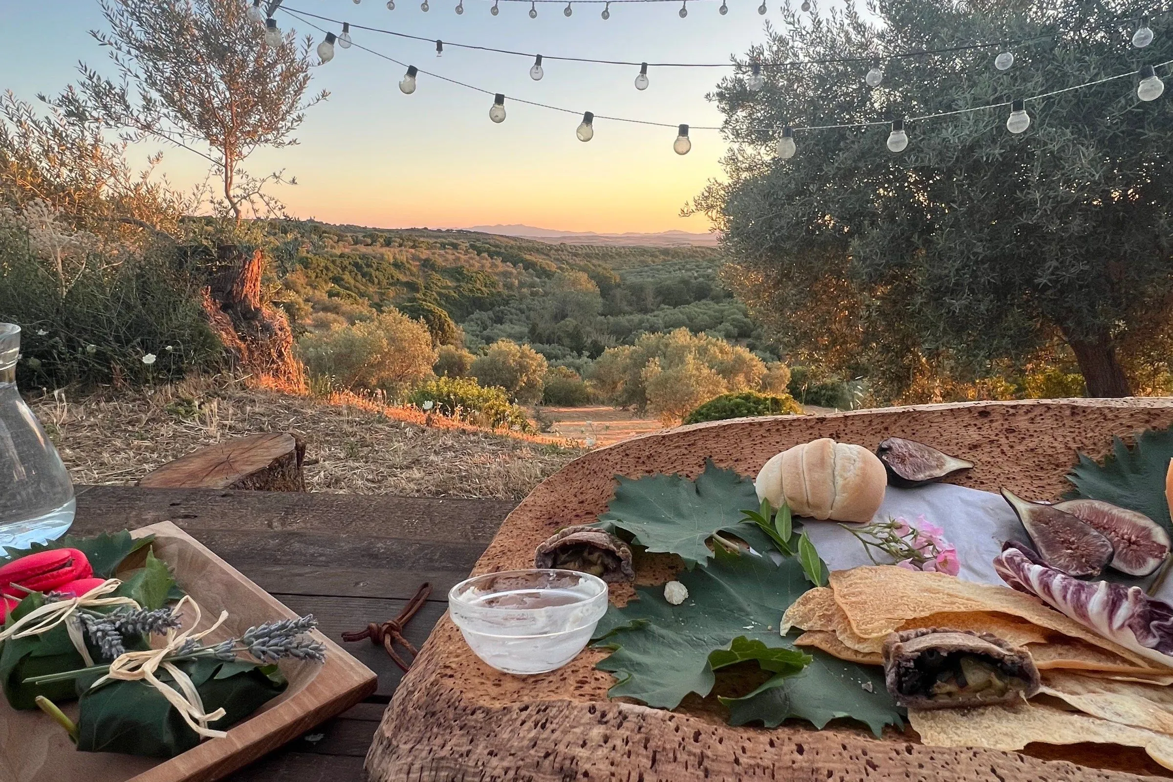 An inviting outdoor dining setup featuring a rustic table with food and a drink bowl, set against a stunning sunset landscape with rolling hills, olive trees, and string lights overhead.